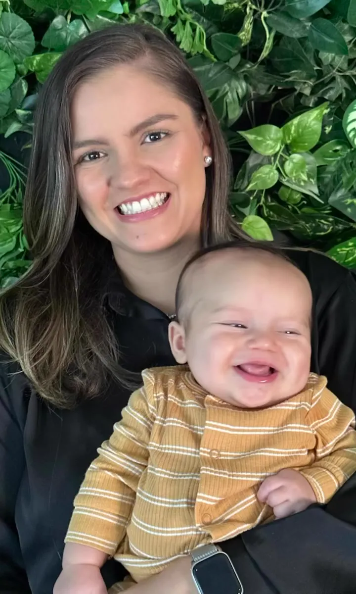 Mariana sorrindo com o filho no colo, em frente a uma parede de folhagem